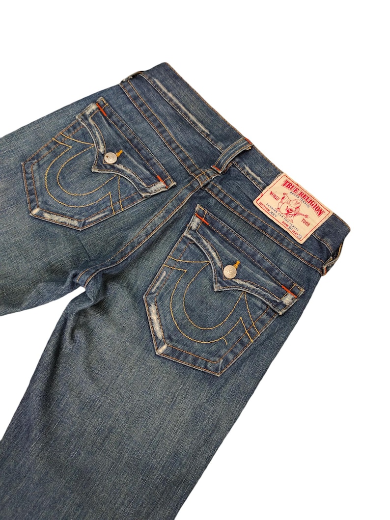 May include: A pair of blue denim jeans with a distressed look. The back pockets have a signature True Religion horseshoe design with stitching in orange and yellow thread. The jeans have a red leather patch with the True Religion logo and the words "True Religion Brand Jeans" printed on it.