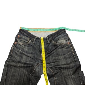 Op de afbeelding: Donkerblauwe denim jeans met een rechte pijp. Een gele meetlint toont de taillemaat. De jeans heeft een klassieke vijf-zakken-stijl en een knoop- en ritssluiting. De taille meet ongeveer 40 cm.