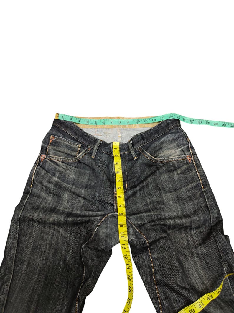Op de afbeelding: Donkerblauwe denim jeans met een rechte pijp. Een gele meetlint toont de taillemaat. De jeans heeft een klassieke vijf-zakken-stijl en een knoop- en ritssluiting. De taille meet ongeveer 40 cm.
