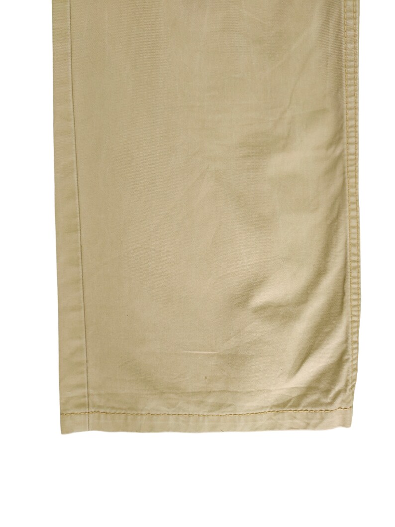 Puede incluir: Primer plano del dobladillo inferior de un pantal&oacute;n caqui. La tela es de algod&oacute;n tejido liso con una textura sutil. El dobladillo est&aacute; acabado con una doble fila de costuras.