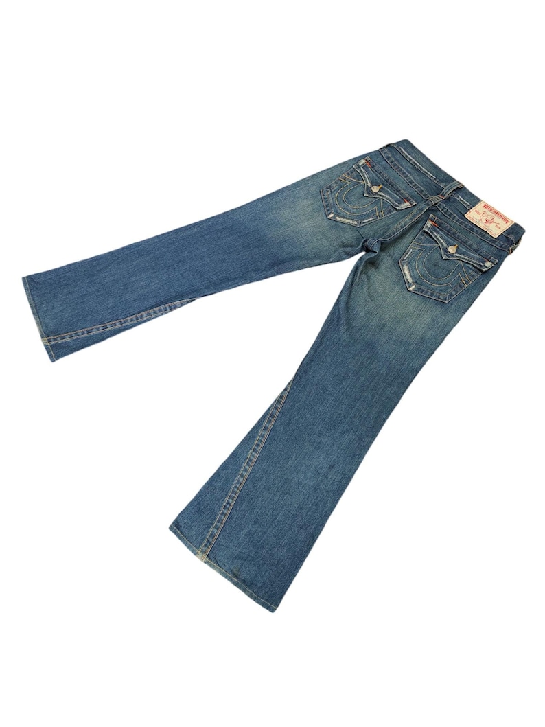 May include: A pair of blue denim jeans with a bootcut leg. The jeans have a light wash and are distressed. The back pockets have a stitched design with the words "True Religion" in white.