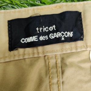 Puede incluir: Un pantal&oacute;n de color beige con una etiqueta negra que dice "tricot COMME des GARCONS".