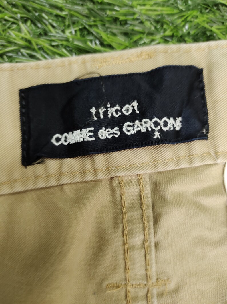 Puede incluir: Un pantal&oacute;n de color beige con una etiqueta negra que dice "tricot COMME des GARCONS".