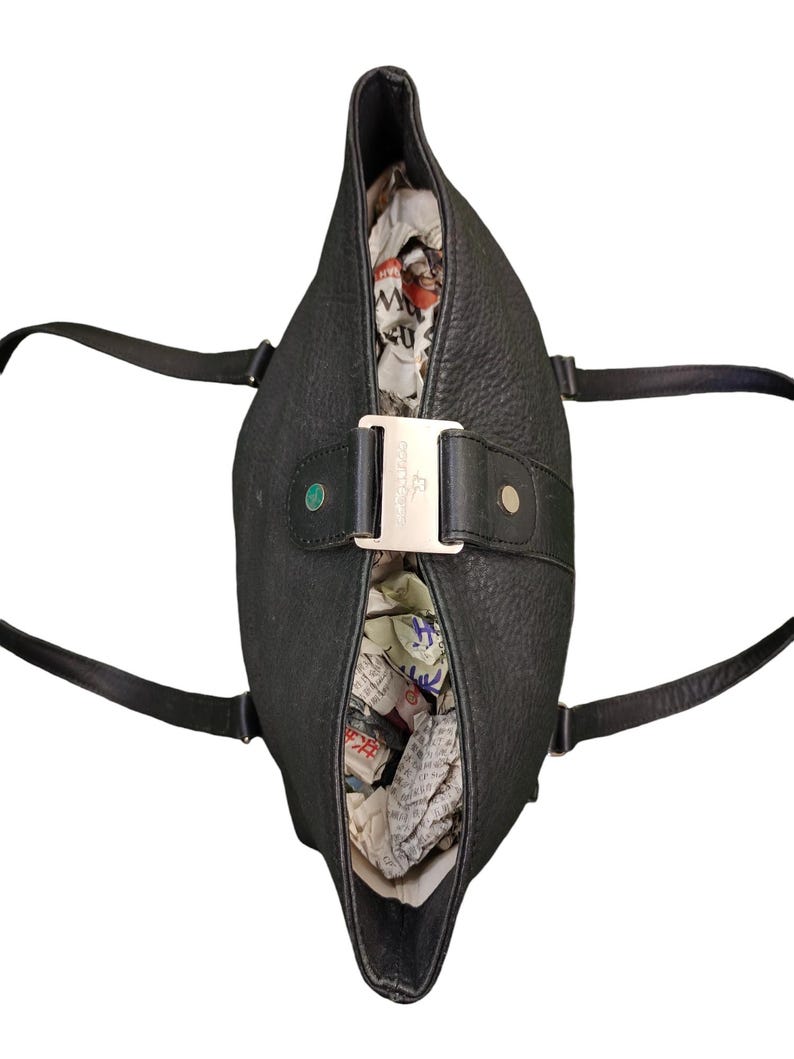 May include: A black leather tote bag with a silver buckle and a small green tag. The bag is open and filled with crumpled paper.