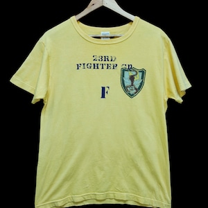 May include: Yellow t-shirt with a blue graphic that says "23rd Fighter GP" and a blue "F" underneath. The graphic also includes a green shield with a scorpion inside.