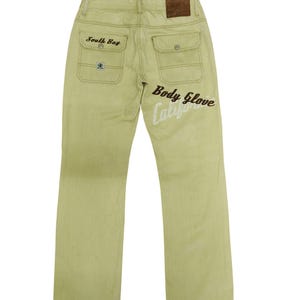 Puede incluir: Vaqueros verde claro con el texto "South Bay" sobre el bolsillo trasero izquierdo y "Body Glove Calif" en la pierna derecha. Los vaqueros tienen dos bolsillos traseros y un parche de cuero marr&oacute;n.