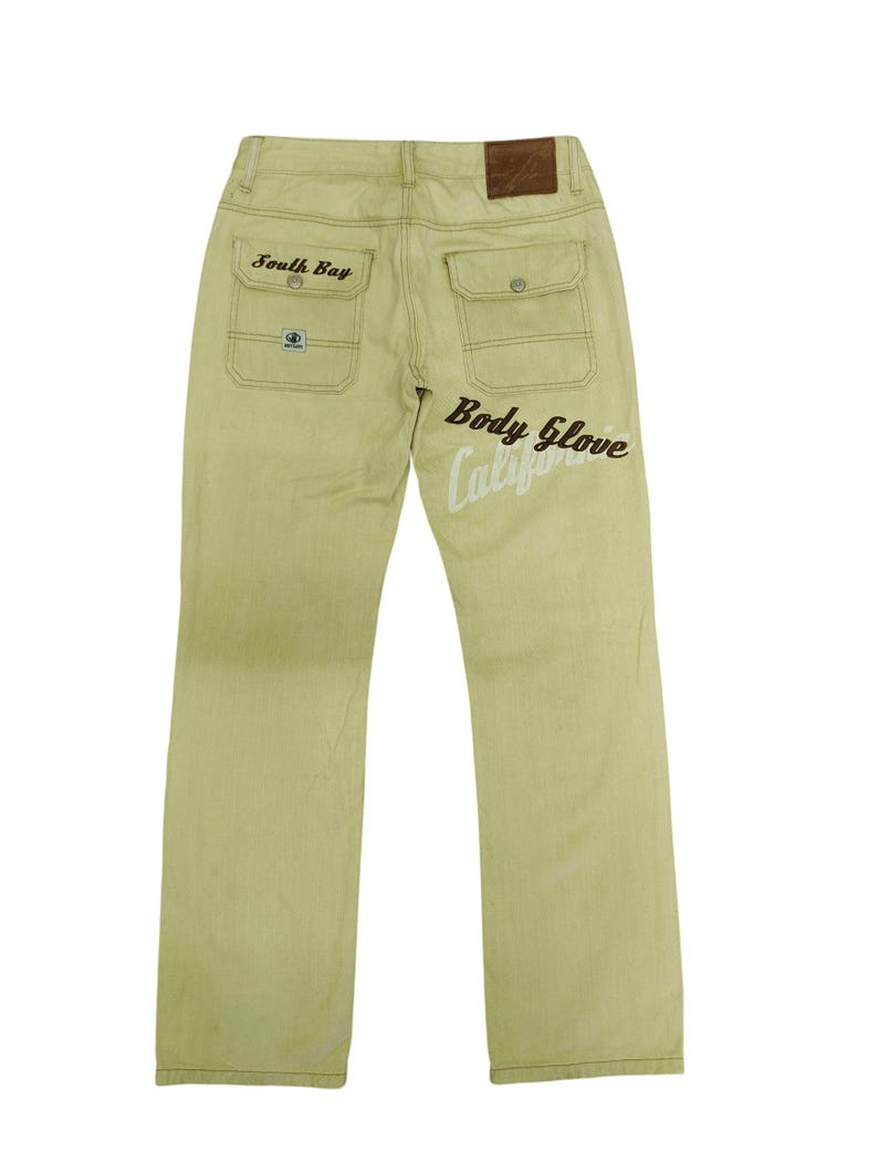 Puede incluir: Vaqueros verde claro con el texto "South Bay" sobre el bolsillo trasero izquierdo y "Body Glove Calif" en la pierna derecha. Los vaqueros tienen dos bolsillos traseros y un parche de cuero marr&oacute;n.