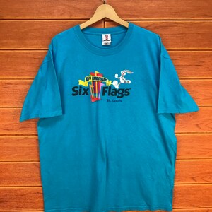 Könnte beinhalten: Türkisfarbenes Six Flags St. Louis 45th Anniversary T-Shirt. Das Shirt zeigt das Six Flags-Logo mit einem Cartoon-Hasen und farbenfrohen Grafiken. Das Shirt hängt an einem Holzbügel vor einem holzgetäfelten Hintergrund.