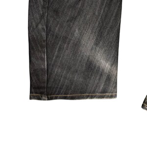 Op de afbeelding: Close-up van een donkergrijze spijkerbroek. De jeans heeft een vervaagde uitstraling met zichtbare stiksels langs de zoom. De stof heeft een textuur en de algehele stijl is casual.