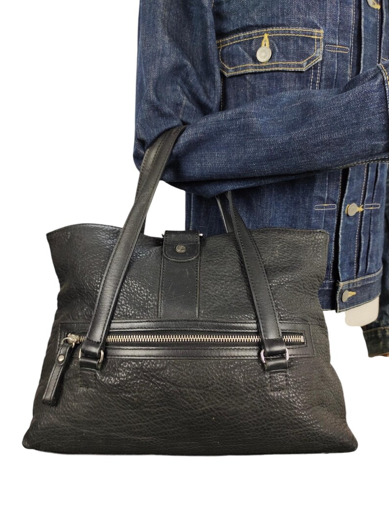 May include: A black leather tote bag with a zipped pocket and two top handles. The bag has a textured surface and a silver zip.