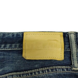 May include: Close-up of the back of blue denim jeans. A light tan leather patch is stitched onto the waistband. The jeans have white stitching and a faded, worn appearance.