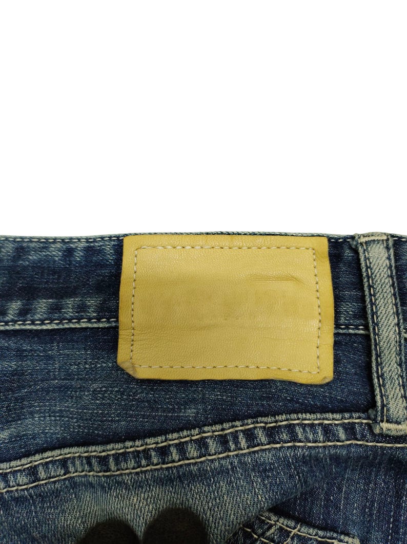 May include: Close-up of the back of blue denim jeans. A light tan leather patch is stitched onto the waistband. The jeans have white stitching and a faded, worn appearance.