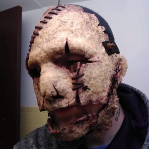 May include: A gruesome, textured mask with a stitched-together appearance. The mask is primarily beige with dark brown and red accents, featuring visible stitches and simulated wounds. The top of the mask has a dark blue section. A horror-themed costume accessory.
