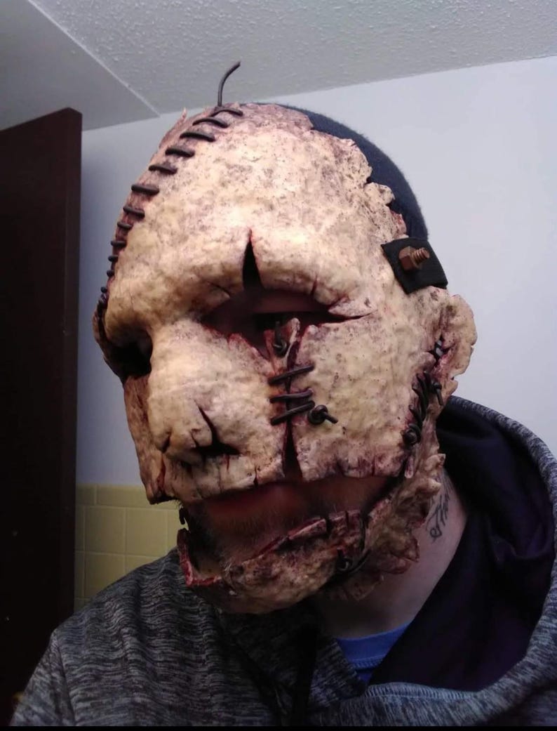 May include: A gruesome, textured mask with a stitched-together appearance. The mask is primarily beige with dark brown and red accents, featuring visible stitches and simulated wounds. The top of the mask has a dark blue section. A horror-themed costume accessory.