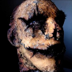 May include: A grotesque, weathered mask with a stitched-up appearance. The mask is primarily tan with dark, jagged cuts around the eyes and mouth, and stitched details. The overall effect is unsettling and horror-themed.