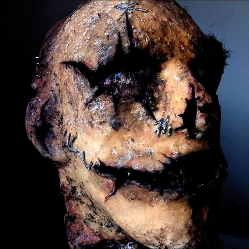 May include: A grotesque, weathered mask with a stitched-up appearance. The mask is primarily tan with dark, jagged cuts around the eyes and mouth, and stitched details. The overall effect is unsettling and horror-themed.