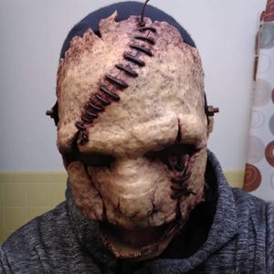 May include: A grotesque mask with a stitched-together appearance, featuring a pale, textured surface with dark red accents. The mask has a stitched seam running down the side and is worn over a dark blue beanie and a gray hoodie.