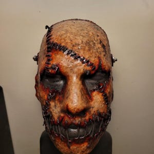 May include: A grotesque mask with a stitched-together appearance. The mask features a textured, tan-colored surface with dark stitching and red accents around the mouth and eyes. The mouth is sewn shut, and the eyes are black.