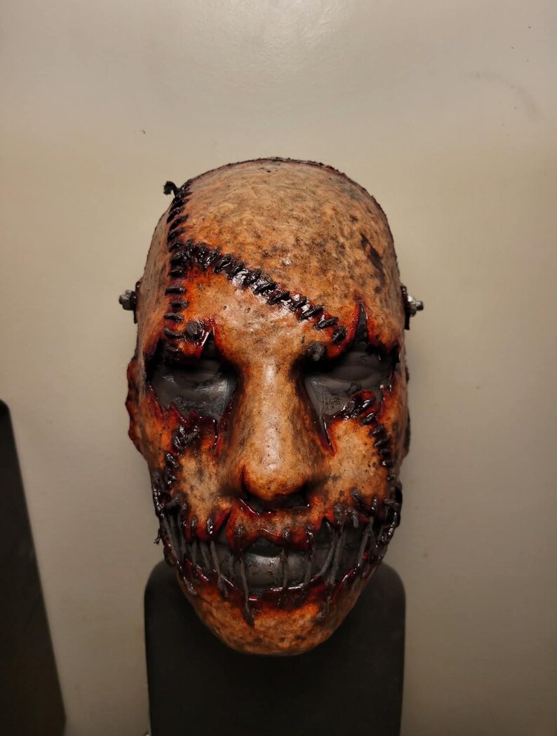 May include: A grotesque mask with a stitched-together appearance. The mask features a textured, tan-colored surface with dark stitching and red accents around the mouth and eyes. The mouth is sewn shut, and the eyes are black.