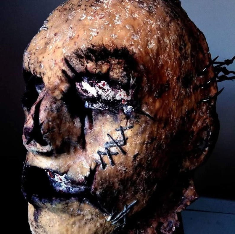 May include: A gruesome, weathered mask with a stitched-up appearance. The mask is primarily brown with dark patches and visible stitches across the face. The eyes are dark and hollow, and the overall effect is unsettling.