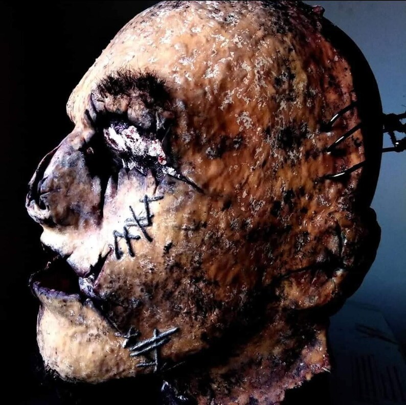 May include: A gruesome, textured mask with a stitched-up mouth and face. The mask is a mix of brown and tan, with dark spots and metal staples. The overall effect is a disturbing, horror-themed design.