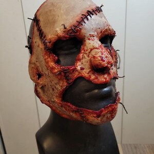May include: A gruesome, realistic mask with a stitched-up design. The mask features a pale, flesh-toned surface with visible stitches and blood-like splatters. The lower face is black, and the mask is mounted on a black mannequin head.