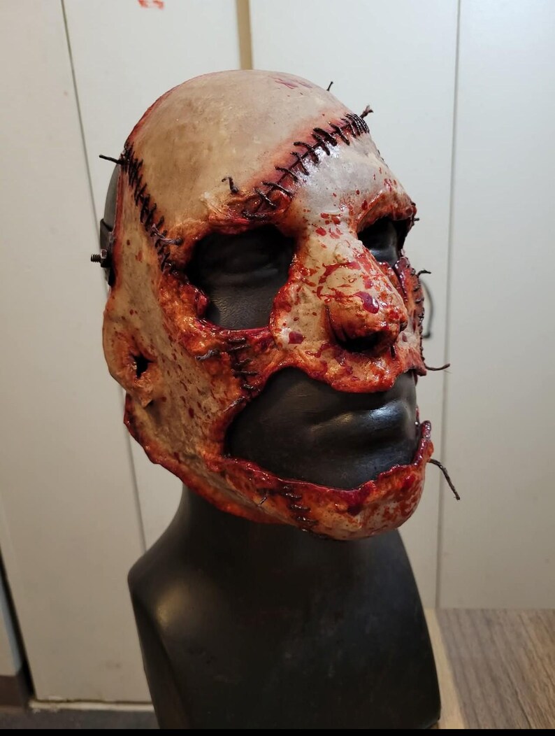 May include: A gruesome, realistic mask with a stitched-up design. The mask features a pale, flesh-toned surface with visible stitches and blood-like splatters. The lower face is black, and the mask is mounted on a black mannequin head.