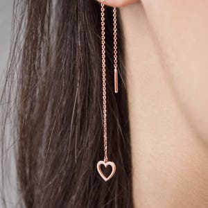 Worn by a model, A threader earring with a dangling heart outline on one end in rose gold.