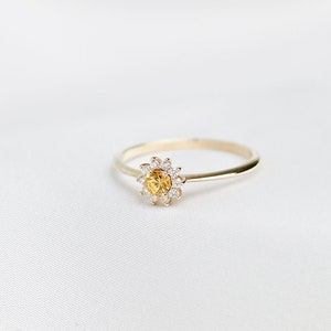 Könnte beinhalten: Ein goldener Ring mit einem gelben Edelstein, der von kleineren weißen Edelsteinen umgeben ist. Der Ring liegt auf einem weißen Stoffhintergrund.