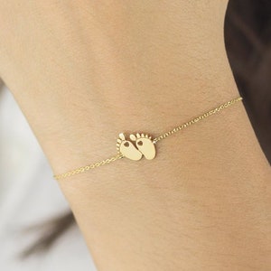 Worn by a model is a cute, yellow gold, tiny baby feet charm with a heart in each one and an adjustable chain surrounding it.