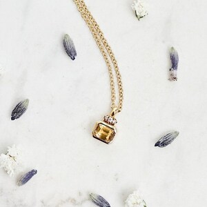 May include: A delicate gold chain necklace with a small, square-shaped pendant featuring a yellow gemstone. The pendant is set in a gold frame and has a small, clear gemstone on top. The necklace is photographed on a white marble surface with small white flowers and purple flower buds scattered around it.
