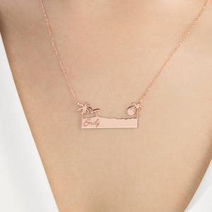 A rose gold pendant necklace with a bar that resembles a beach with an engravable message, a palm tree on each side an umbrella on the left, and the sun on the right is shown worn by a model.