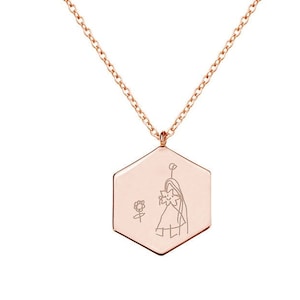 Op de afbeelding: Roségouden ketting met een zeshoekige hanger. De hanger heeft een lijntekening van een persoon die een bloem vasthoudt. De ketting en hanger zijn roségoudkleurig. Het ontwerp is eenvoudig en artistiek. Een delicaat sieraad.