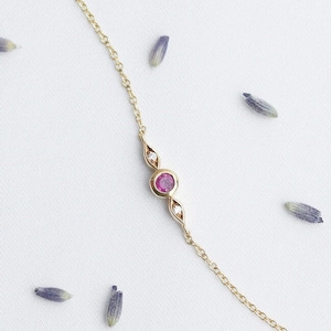 Può includere: Una delicata collana a catena dorata con una piccola pietra preziosa rosa circondata da due piccole pietre preziose chiare.