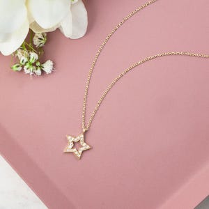 Laid down on a pink display piece and with flowers next to it, a yellow gold necklace with a special star outline charm and natural white diamonds embedded in it.