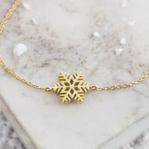 Puede incluir: Un delicado collar de cadena dorada con un colgante de copo de nieve. El collar está sobre una superficie de mármol blanco.
