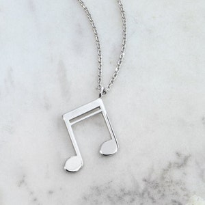 May include: A silver chain necklace with a silver musical note pendant. The necklace is on a white marble surface with white flowers.