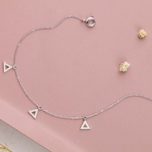 Könnte beinhalten: Ein filigranes silbernes Fußkettchen mit drei Dreiecksanhängern. Das Fußkettchen liegt auf einer rosa Oberfläche, mit kleinen getrockneten weißen Blüten. Schmuck ist schlicht und elegant.
