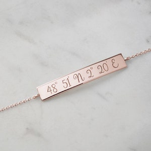 A rose gold bracelet with an engravable horizontal curved bar with coordinates attached to a chain that has its jump rings soldered and a spring-ring clasp is shown on a marble surface.