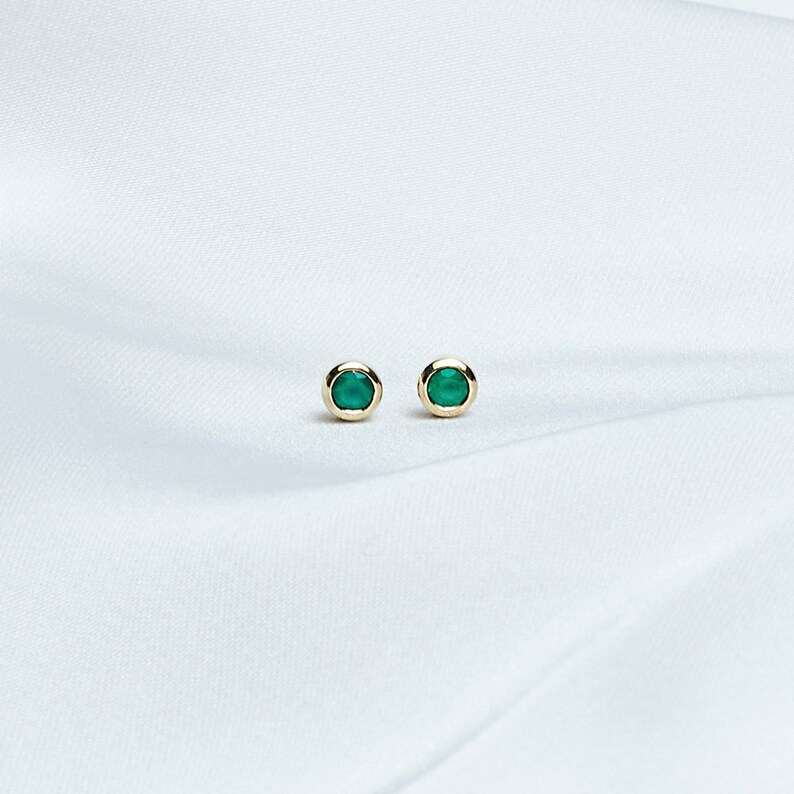 A Beautiful set of yellow gold stud earrings with a tiny round green agate gemstone laid down on a white sheet.