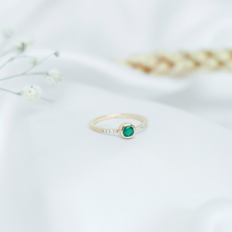 Laid down on a white sheet with flowers around it, a beautiful yellow gold ring with a round-shaped green agate gemstone and five tiny diamonds on each side of the stone, around its band.