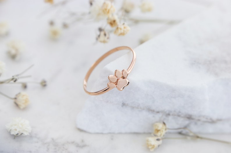 A paw print band ring with a distinct separation between the pads in rose gold laid down a piece of marble with pedals spread across the picture.