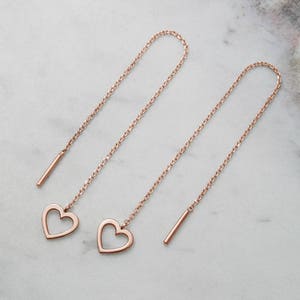 Laid down on a marble piece, A threader earring with a dangling heart outline on one end in rose gold.