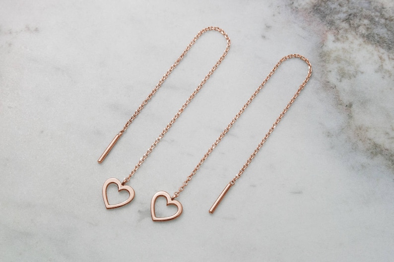 Laid down on a marble piece, A threader earring with a dangling heart outline on one end in rose gold.