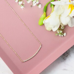 A minimalist style yellow gold necklace with a thin cable chain and a curved bar with natural white diamonds embedded in it. The piece is beautifully shown on a pink display with flowers next to it.