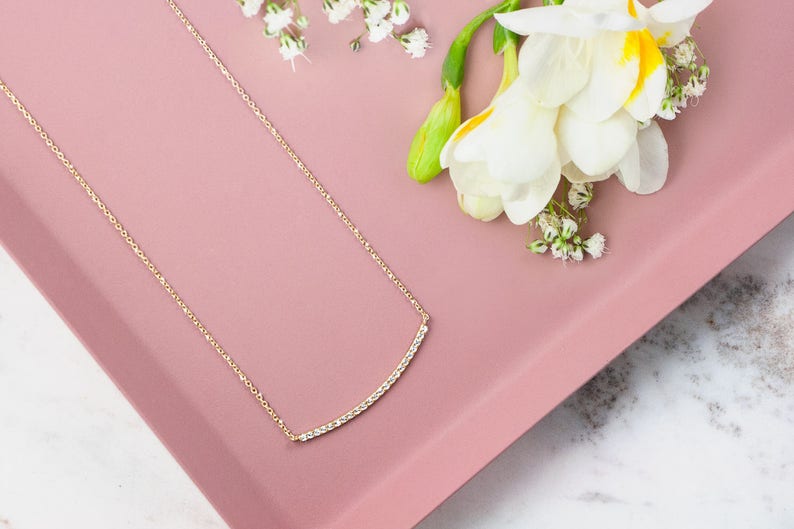 A minimalist style yellow gold necklace with a thin cable chain and a curved bar with natural white diamonds embedded in it. The piece is beautifully shown on a pink display with flowers next to it.