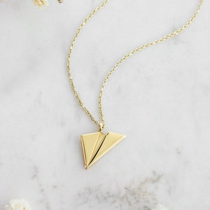 A unique yellow gold necklace with a tiny flat paper plane charm attached to it is shown laid down on a marble surface with flowers surrounding it.
