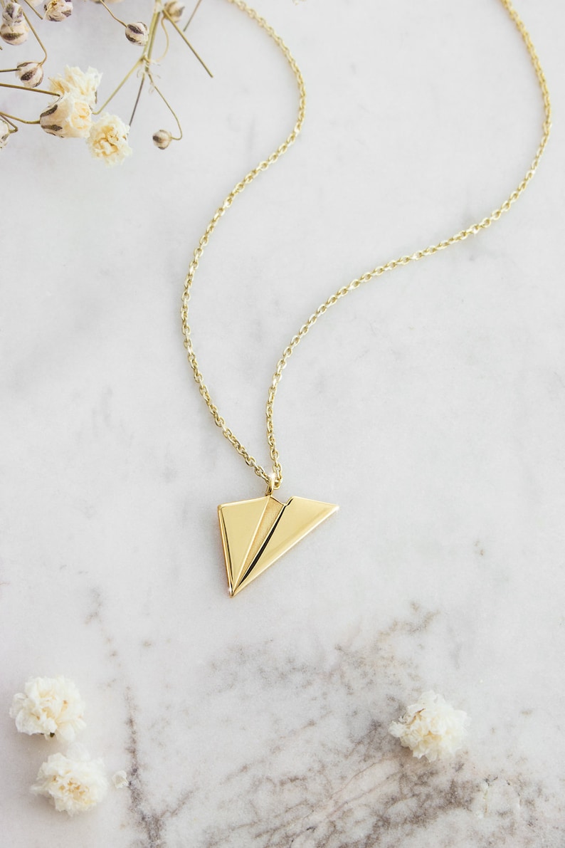 A unique yellow gold necklace with a tiny flat paper plane charm attached to it is shown laid down on a marble surface with flowers surrounding it.