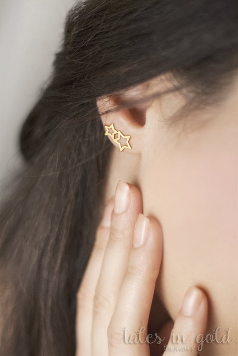 A pair of different-sized two-star outline stud earrings in yellow gold is shown worn by a model.