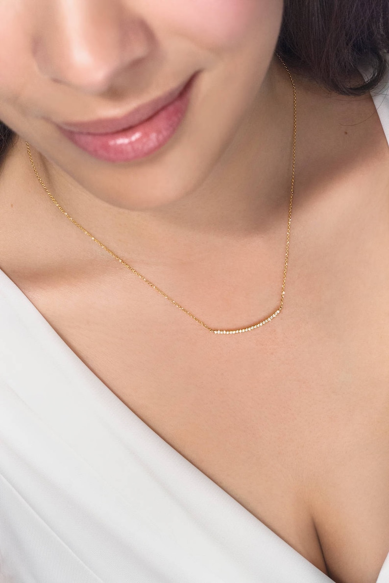A minimalist style yellow gold necklace with a thin cable chain and a curved bar with natural white diamonds embedded on it worn by a model.